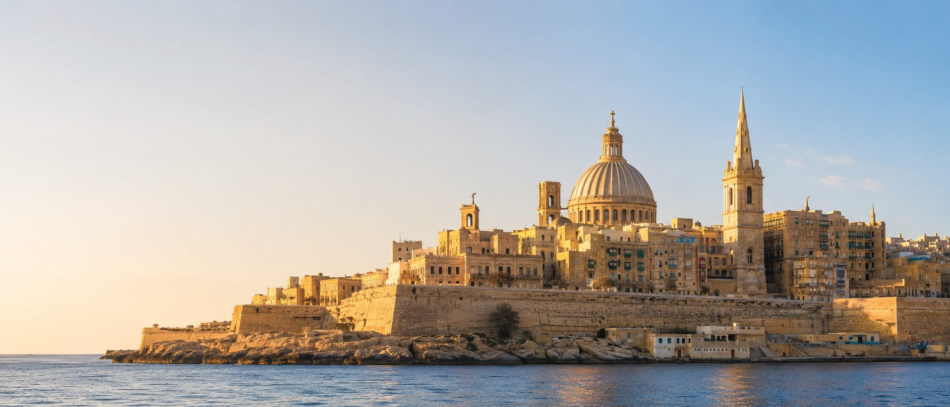 The limestone skyline of Valletta at golden hour, with the Carmelite dome and St Paul's spire rising above the fortress walls and the sea